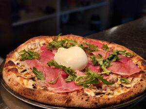 Delicious gourmet pizza topped with fresh prosciutto, arugula, and a dollop of creamy burrata cheese, highlighting the vibrant flavors of Amalfi Bar & Pizzeria in Seminyak, Bali.