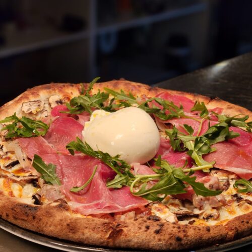 Delicious gourmet pizza topped with fresh prosciutto, arugula, and a dollop of creamy burrata cheese, highlighting the vibrant flavors of Amalfi Bar & Pizzeria in Seminyak, Bali.